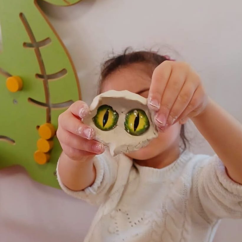 Ein Kind hält mit stilisierten reptilienartigen Augen vesehenes Knetstück in die Kamera.