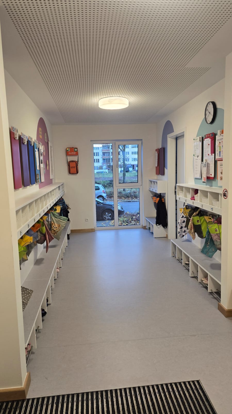 Flur mit Regalen und bunten Bildern, Eingangstür im Hintergrund.