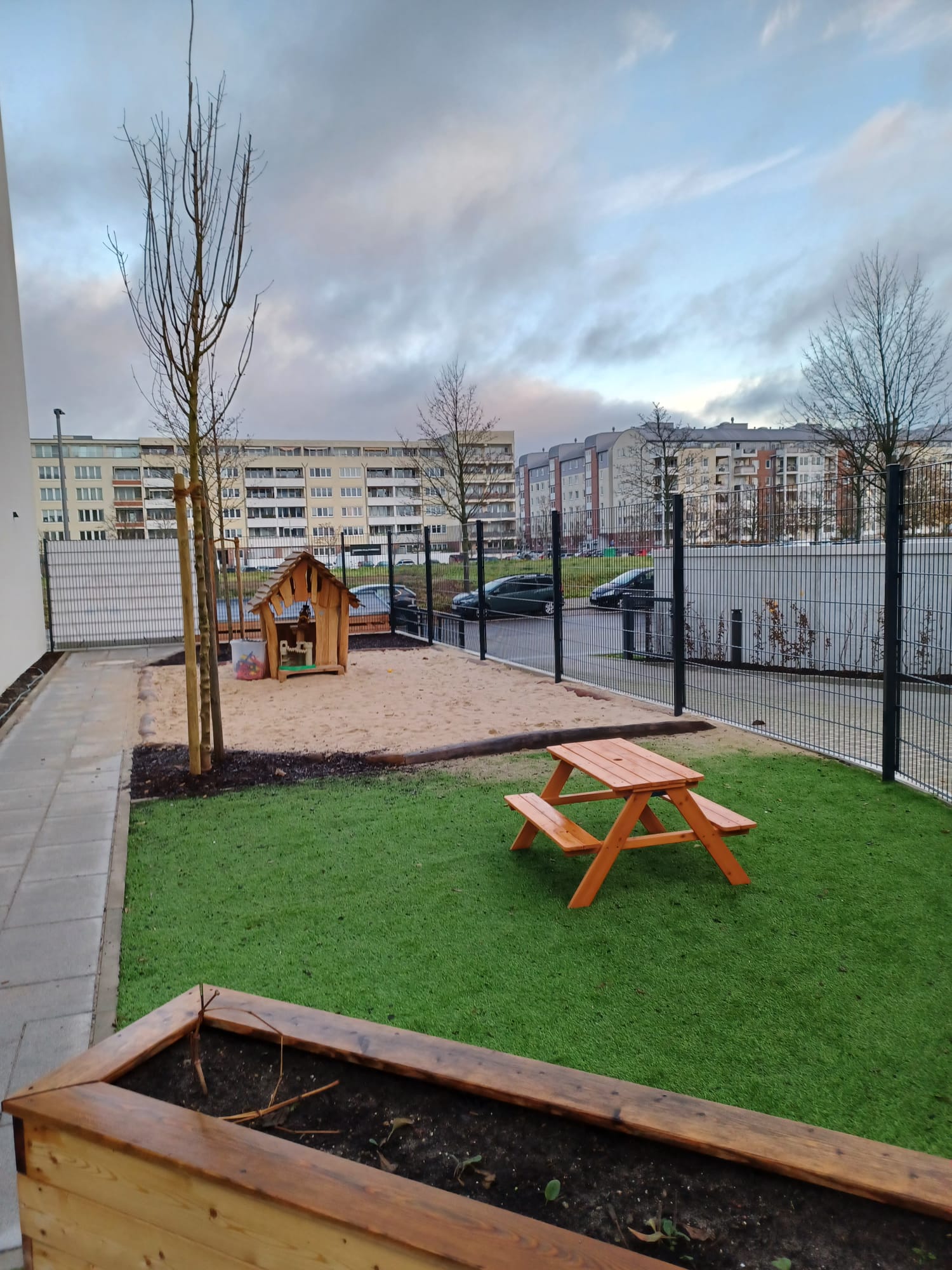 Spielplatz mit Holzschaukel und Picknicktisch auf Grasfläche. Hintergrund mit Gebäuden.