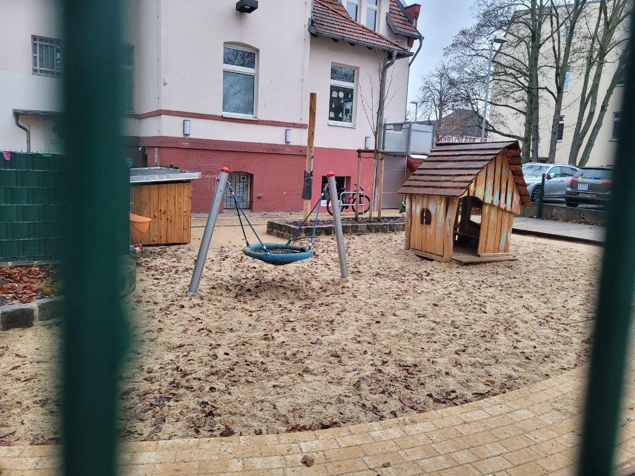 Spielplatz mit Schaukeln, Holzhaus und sandigem Boden, umgeben von einem Zaun.