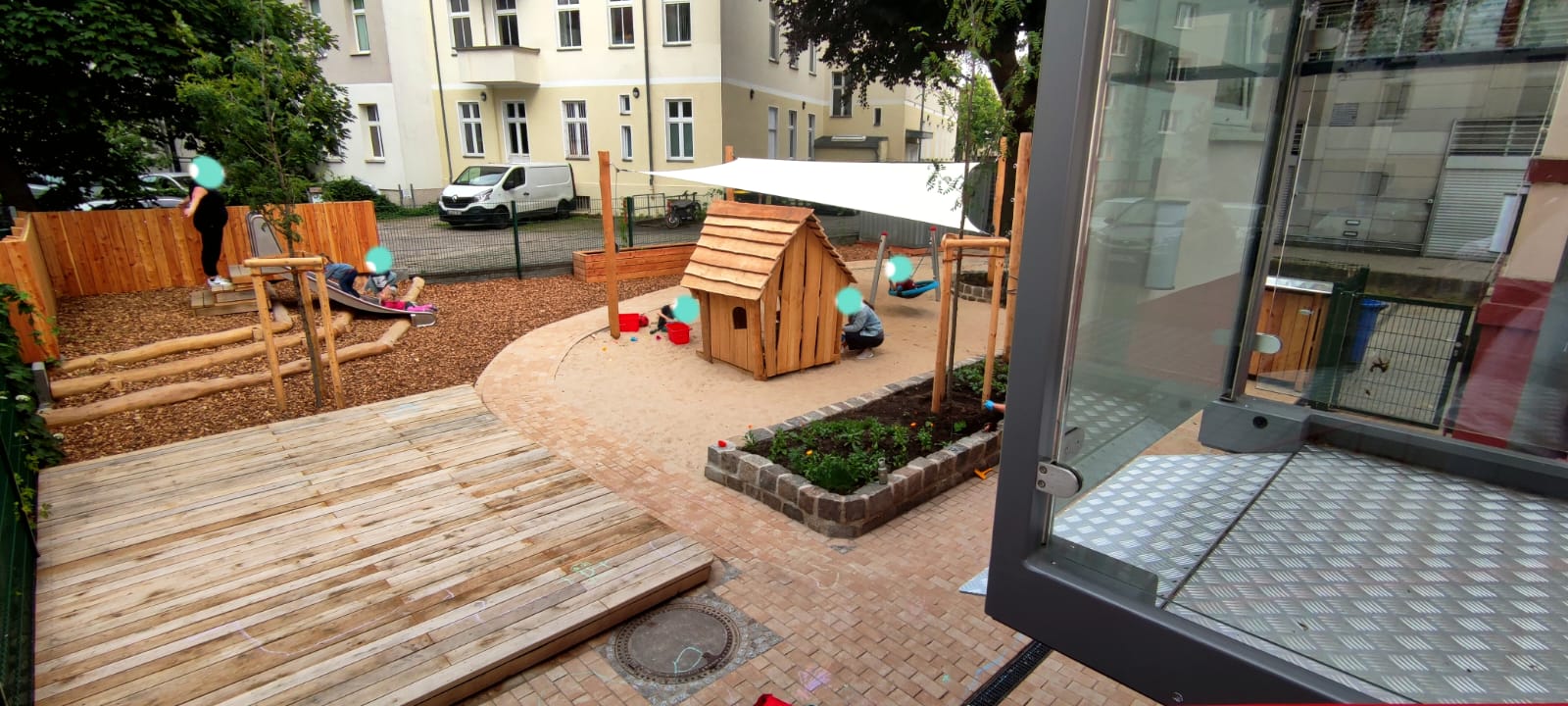 Spielplatz mit Holzspielhaus, Sandfläche und Grünflächen, umgeben von Gebäuden.