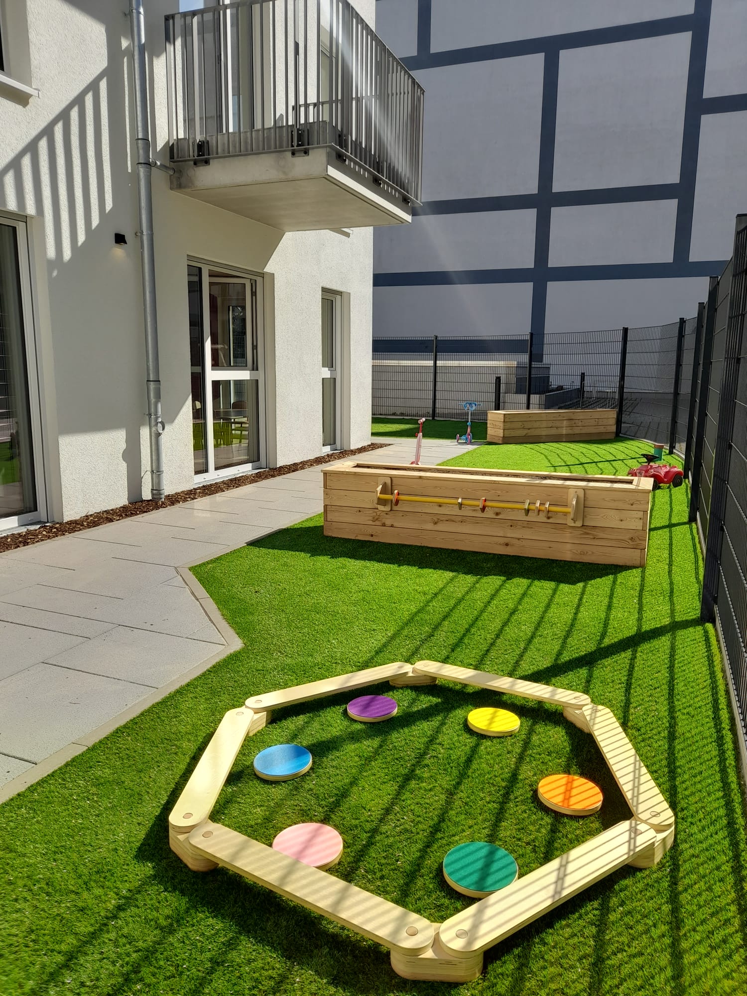 Spielplatz mit Rasen, Holzspielgeräten und bunten Steinflächen.