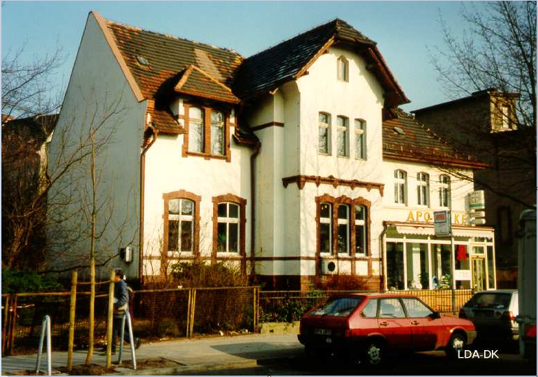 Helles Gebäude im historischen Stil mit roten Details und angrenzendem Parkplatz.