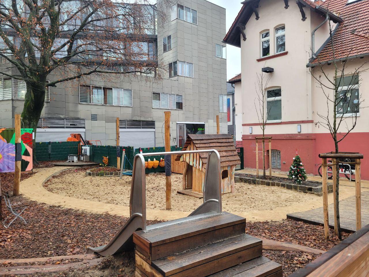 Spielplatz mit Holzspielgeräten, Sandfläche und angrenzendem Wohngebäude.